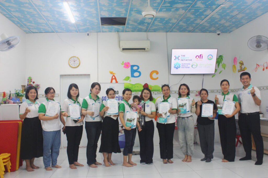 Group of adults standing in a bright classroom, each holding a certificate and posing for a photo smile.