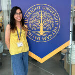 Smiling woman in a yellow top with a lanyard stands beside a large blue banner featuring a golden tree emblem and circular Fulbright University text.