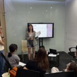 Woman presenting in a glass-walled conference room, speaking to a small audience gathered around.