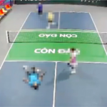 Top-down view of a doubles tennis match on a blue court with a green central service area and a net; four players are moving around near the net.