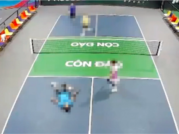 Top-down view of a doubles tennis match on a blue court with a green central service area and a net; four players are moving around near the net.