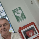 Older man in white shirt standing beside an AED sign and a red HeartStart defibrillator cabinet on a wall behind him