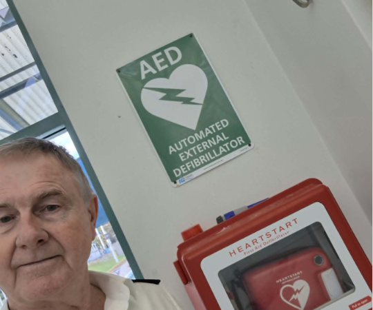 Older man in white shirt standing beside an AED sign and a red HeartStart defibrillator cabinet on a wall behind him