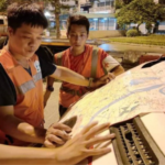 Three workers in orange reflective vests study a large city map spread over the hood of a vehicle at night on a street.