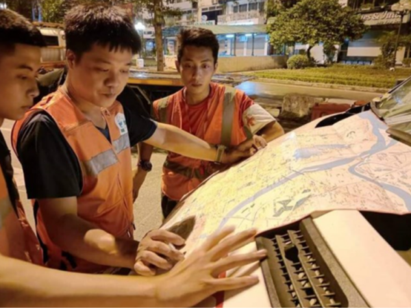 Three workers in orange reflective vests study a large city map spread over the hood of a vehicle at night on a street.