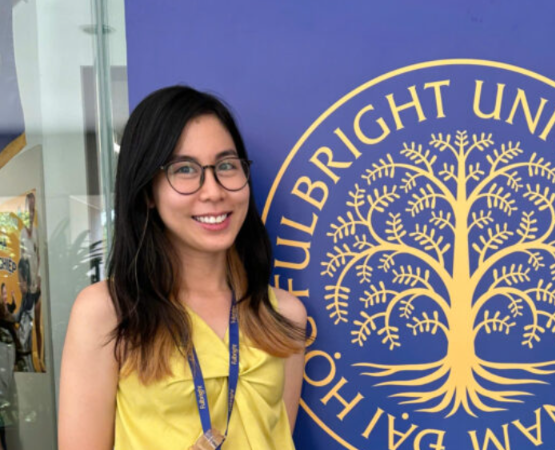 Smiling woman with glasses in a yellow top stands beside a blue banner featuring a gold tree emblem and text, wearing a blue lanyard.