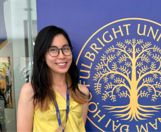 Smiling woman with glasses in a yellow top stands beside a blue banner featuring a gold tree emblem and text, wearing a blue lanyard.