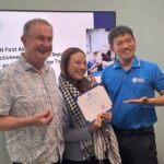 Three smiling people pose for a photo: a man in a patterned shirt, a woman holding a certificate, and a man in a blue polo presenting it in front of a presentation screen.
