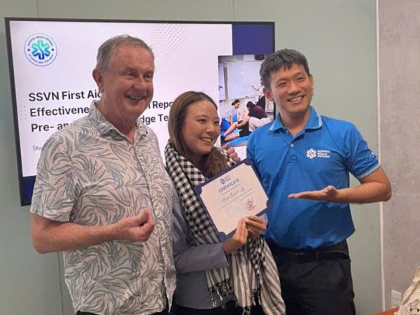 Three smiling people pose for a photo: a man in a patterned shirt, a woman holding a certificate, and a man in a blue polo presenting it in front of a presentation screen.